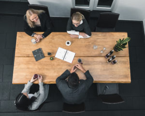 Corporate Resilience Management – 3-core 19 Vier Personen sitzen um einen Holztisch mit Notizbüchern, Kaffeebechern und Wasserflaschen und führen eine lebhafte Diskussion über die Krisenvorsorge. Eine Topfpflanze begrünt die Szene. Das Foto ist von oben aufgenommen.
