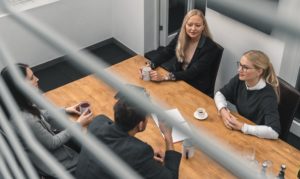 Behördliche Krisenstabsübung 5 Vier Personen sitzen um einen Holztisch in einem Büro und besprechen ein neues Konzept. Drei halten Tassen in der Hand und einer hat eine Tasse Kaffee. Das Foto ist durch die Jalousien aufgenommen, auf dem Tisch stehen Papiere und Wassergläser.