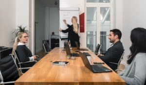 Four people are in a modern conference room. Three are seated at a wooden table with laptops, while one person stands at a flipchart, writing key points on construction protection. The atmosphere appears professional and focused.
