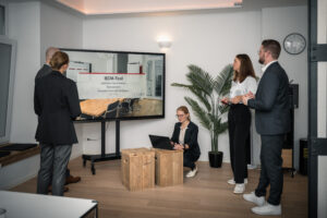 Blog 11 A group of five people in business attire are having a meeting in a modern office. One person is presenting a slide titled “BCM-Tool” on a large screen, discussing strategies for emerging market entry, while another sits with a laptop on wooden crates.