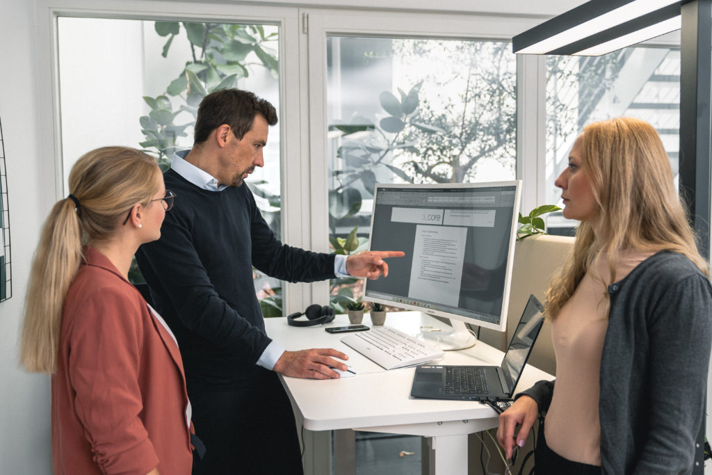 Three people stand around a computer monitor in an office, discussing the document displayed on the screen. One person points at the monitor while the others listen attentively, highlighting their strong focus on corporate security.