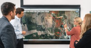 A Crisis Team Training session unfolds as four people gather around a large screen displaying a satellite map; one woman draws red markings and arrows on the map while the others observe and discuss.