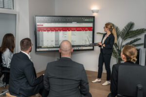 Blog 9 A woman in business attire presents data on a large screen to four colleagues during a Security Awareness Kampagne in an office setting, with a potted plant in the background.