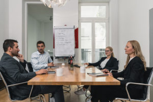 Four people in business attire sit around a conference table in a modern office, discussing notes for a Security Awareness Kampagne. Notebooks and coffee mugs are on the table, and a flipchart with ideas stands near the window.