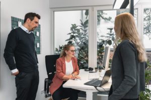 Three people in an office, two standing and one sitting at a desk with a computer, appear to be discussing a Security Awareness Kampagne. Large windows and plants add to the bright, modern workspace.