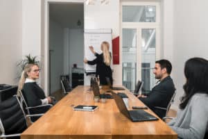 Blog 7 Four people are in a modern meeting room. One woman stands at a flip chart, presenting certification updates to three colleagues seated at a long wooden table with laptops, notebooks, and drinks.