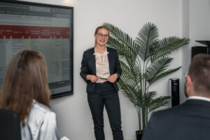 A woman in business attire stands and presents with the BCM-Tool displayed on a large screen to two seated colleagues in an office, with a tall potted plant beside her.