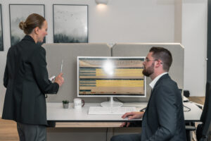 Blog 9 Two people in business attire discuss data related to business impact analyses displayed on a computer monitor at a modern office desk, with documents, a keyboard, and a coffee mug visible. One person is standing, the other is seated.