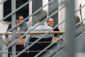 Blog 11 Four people have a meeting around a wooden table in a modern office, conducting risk analyses. Two men and one woman are visible, with one woman smiling; the image is partially obscured by metal railings in the foreground.