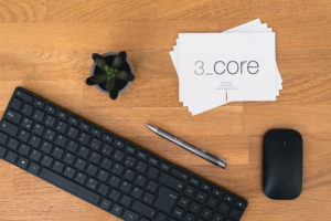 A black computer keyboard, silver pen, and black wireless mouse sit on a wooden desk beside a small potted succulent and a stack of cards labeled 3_core smart design management with notes on risk analyses.