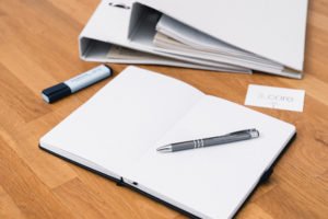 Behördliche Krisenstabsübung 4 Ein aufgeschlagenes Notizbuch mit einem Stift liegt auf einem hölzernen Schreibtisch. Daneben liegen ein blauer Textmarker, zwei weiße Ringbücher mit Unterlagen über Prozesse im Kontext BCM und eine Visitenkarte mit der Aufschrift 3_core.