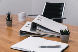 A wooden desk with two open white binders filled with papers from recent risk analyses, a notebook, a pen, two small potted plants, a mug, and an empty black office chair in the background.
