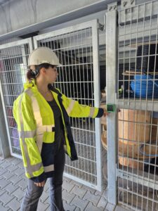 A person wearing a hard hat, safety glasses, and a high-visibility jacket is conducting a pentest by pressing buttons on a keypad lock attached to a metal gate in an industrial storage area.
