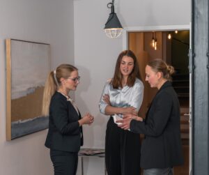 Three women in business attire stand in a modern office hallway of a public security institution, engaged in conversation. One gestures while holding papers, while the others listen attentively. A large painting and hanging light fixture are visible.