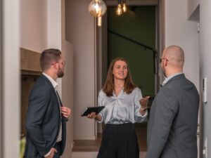 Eine Frau in einer hellen Bluse und dunklen Hose spricht in einem modernen Büro mit zwei Männern in Anzügen, während sie ein Notizbuch in der Hand hält und gemeinsam über Krisenmanagement für Unternehmen sprechen.