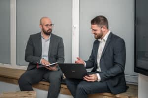 3-core im Interview mit Welt TV: Öffentliche Sicherheit unter Druck 5 Zwei Männer in Anzügen sitzen auf einer Holzbank in einem Büro und besprechen ihre Arbeit. Der eine hält ein Notizbuch und einen Stift in der Hand, während der andere mit einem aufgeklappten Laptop gestikuliert, während sie sich auf ein Track-and-Trace-Projekt konzentrieren, wobei beide in ein Gespräch vertieft zu sein scheinen.