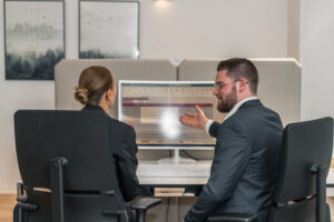 Two people in business attire sit at an office desk, reviewing spreadsheets related to emergency planning. One gestures toward the screen while explaining details to the other. Office decor and framed art are visible in the background.