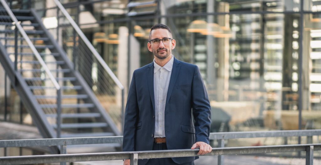 A man in a navy blue suit and glasses stands outdoors, holding a 3-core railing. Behind him are metal stairs and large glass windows reflecting modern office lights.