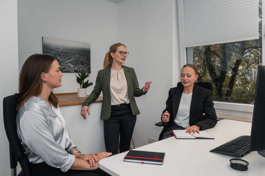 Three women in business attire meet in a modern office. One woman stands and speaks, while the other two sit at a desk with notebooks, listening attentively. A computer, plant, and large window are in the background.