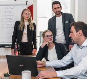 Corporate Resilience Management – 3-core 19 Vier Personen in Geschäftskleidung sind in einem Büro um einen Tisch mit einem Laptop und Dokumenten versammelt. Zwei stehen an einem Flipchart mit Notizen, während zwei im Sitzen die Risk Governance diskutieren.