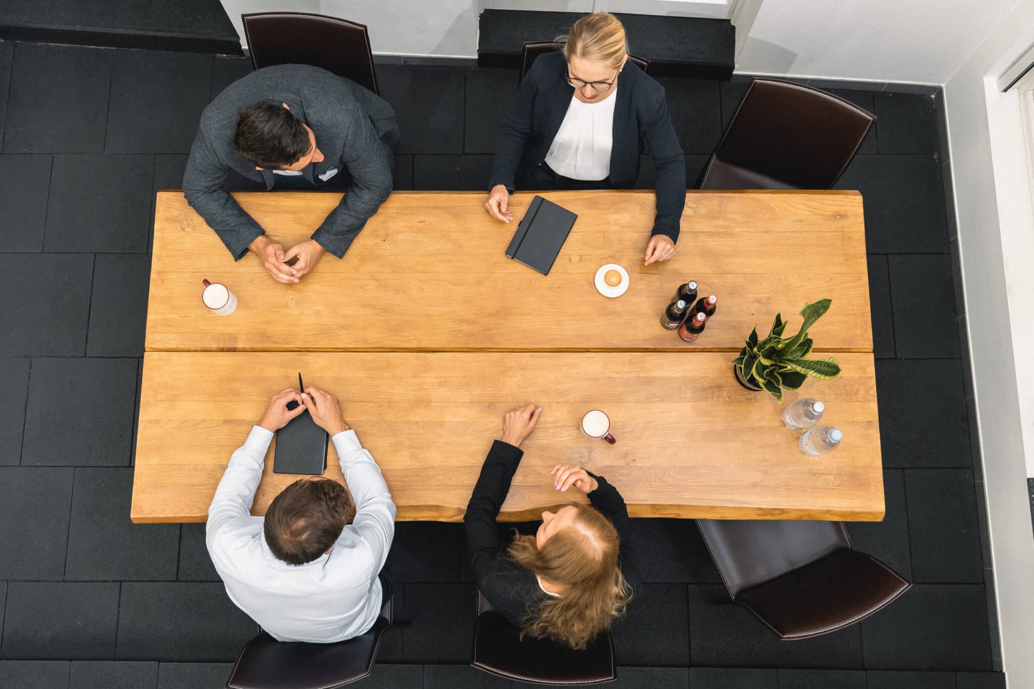 Four people in business attire sit around a rectangular wooden table in a modern office, viewed from above, discussing KRITIS implementation. The table holds notebooks, drinks, coffee cups, a potted plant, and bottled water.