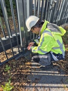 A person wearing a white hard hat and a bright yellow safety jacket kneels beside a metal fence, working with tools on the gate—demonstrating civil engineering practices near fallen leaves and gravel.