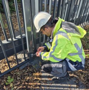 Schwarzstartfähig und geschützt: Sicherheitsplanung für Kraftwerksstandorte 4 Eine Person mit weißem Schutzhelm und Warnweste kniet auf dem Boden neben einem Metallzaun und arbeitet mit Werkzeugen in der Nähe des Torsockels - ein Beispiel für das Sicherheitskonzept Baustelle in Aktion. Auf dem Boden sind trockene Blätter und Schotter zu sehen.
