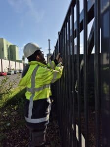 Blog 11 A person wearing a white safety helmet and a bright yellow reflective jacket is working on or inspecting a tall metal fence outdoors, possibly as part of plant protection efforts, with industrial buildings and a chimney in the background.