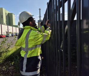 KRITIS-Dachgesetz 2026: Anforderungen, Pflichten und Audit-Readiness 3 Eine Person, die eine Warnweste und einen weißen Schutzhelm trägt, steht an einem hohen Metallzaun und inspiziert diesen auf einem schwarzstartfähigen Industriegelände, mit Gebäuden und einem Schornstein im Hintergrund.