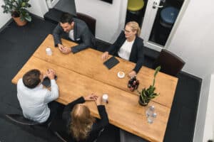 Blog 7 Four people in business attire sit around a wooden table discussing strategies. Drinks, a tablet, bottles, and a plant are on the table. The modern office setting hints at a Consulting Expert für KRITIS leading the meeting, viewed from above.