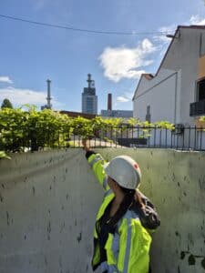 From an Exceeded threshold to a Security Concept 6 A woman wearing a helmet and a high-visibility vest points to a security fence as a visible security measure on a site in clear, sunny weather. The scene illustrates a Security Requirements Analysis in practice.