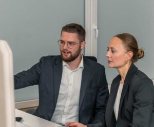 Corporate Resilience Management – 3-core 19 Zwei professionell gekleidete Personen sitzen an einem Schreibtisch und diskutieren über die NIS2 Umsetzung, während sie auf einen Computerbildschirm schauen. Einer zeigt auf den Bildschirm, während der andere Notizen in ein Notizbuch schreibt. Auf dem Schreibtisch stehen eine kleine Pflanze und ein Becher.