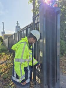 Securing critical infrastructure: 3-core at the PROTEKT conference 4 A person wearing high-visibility clothing is working on a metal fence that forms part of a Security Zone. In the background, a facility operated by an Energy Producer is visible.