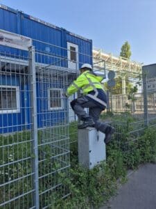 KRITIS Umbrella Act 2026: Requirements, Obligations, and Audit Readiness 3 An employee in high-visibility clothing is installing or inspecting the perimeter protection on a metal fence in front of an industrial building.