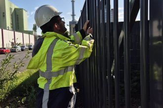A person wearing a white safety helmet and a bright yellow reflective jacket is working on or inspecting a tall metal fence to verify that it meets the required security threshold for perimeter protection, with industrial buildings and a chimney in the background.