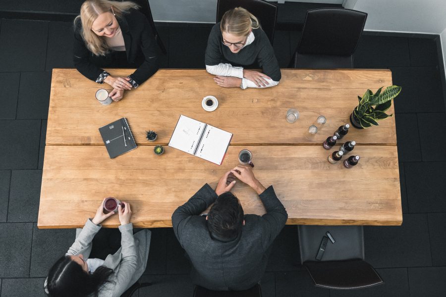 Vier Personen sitzen um einen Holztisch mit Notizbüchern, Kaffeebechern und Wasserflaschen und führen eine lebhafte Diskussion über die Krisenvorsorge. Eine Topfpflanze begrünt die Szene. Das Foto ist von oben aufgenommen.