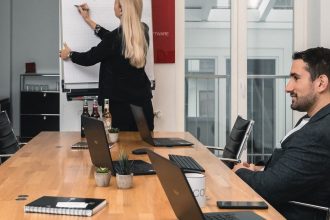 Four people are in a modern conference room. Three are seated at a wooden table with laptops, while one person stands at a flipchart, writing key points on construction protection. The atmosphere appears professional and focused.
