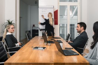 Vier Personen befinden sich in einem Besprechungsraum. Drei sitzen an einem Holztisch mit Laptops, während einer an einem Flipchart steht und Notizen schreibt. Der Raum ist hell, hat große Fenster und ist minimalistisch eingerichtet.
