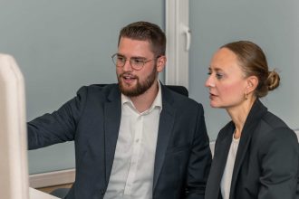 Zwei professionell gekleidete Personen sitzen an einem Schreibtisch und diskutieren über die NIS2 Umsetzung, während sie auf einen Computerbildschirm schauen. Einer zeigt auf den Bildschirm, während der andere Notizen in ein Notizbuch schreibt. Auf dem Schreibtisch stehen eine kleine Pflanze und ein Becher.
