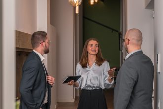 Eine Frau in einer hellen Bluse und dunklen Hose spricht in einem modernen Büro mit zwei Männern in Anzügen, während sie ein Notizbuch in der Hand hält und gemeinsam über Krisenmanagement für Unternehmen sprechen.