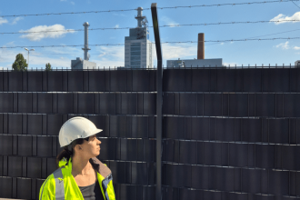 Eine Person, die einen weißen Schutzhelm und eine gelbe Warnweste trägt, steht im Freien in der Nähe eines Zauns, mit Industriegebäuden, die möglicherweise dem Kritis Dachgesetz unterliegen, und einem blauen Himmel mit vereinzelten Wolken im Hintergrund.