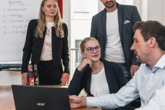 Vier Personen in Geschäftskleidung sind in einem Büro um einen Tisch mit einem Laptop und Dokumenten versammelt. Zwei stehen an einem Flipchart mit Notizen, während zwei im Sitzen die Risk Governance diskutieren.