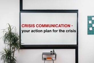 A whiteboard displays: Crisis communication - your action plan for the crisis! Nearby, there’s a potted plant, a small table with items, and a green board featuring papers.