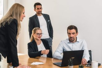 Vier Personen in Geschäftskleidung versammeln sich um einen Holztisch und konzentrieren sich auf einen Laptop, während sie über KRITIS DachG Umsetzung diskutieren. Zwei sitzen, zwei stehen in der Nähe in einem modernen, minimalistisch eingerichteten Büro.